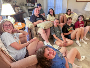 Logan Byrd is one of 10 people posing for a photo in a family room. He sits on a couch with a pillow on his knees, his hands folded in his lap, and smiles at the camera. Two girls sit beside him on the couch, while three girls sit on the floor leaning against it. Another boy sits on a chair behind the couch, and two girls sit on and beside a nearby recliner. One or two additional people are partially visible in the background. Family photos and other household items are visible throughout the room. Everyone is casually dressed in T-shirts and comfortable clothing, wearing socks but no shoes.