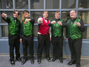Logan Byrd is one of five high school-aged young men posing in front of a wall of square windows, likely in a high school. All five wear black pants and dress shoes with dress shirts, vests, and ties. Logan, standing on the far right, and three of the other young men wear black shirts and vests with a sparkly green looping pattern and matching neckties. The fifth young man wears a white dress shirt with a shiny red vest and red bow tie. All five hold up medals they appear to have recently won, and Logan smiles widely at the camera.