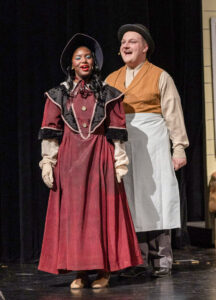 Logan Byrd performs on stage with a young woman, both appearing to be high school students. Logan stands behind her, singing and wearing a vintage-style tan shirt, mustard-yellow vest and pants, white apron, and bowler hat, appearing to portray an old-fashioned shopkeeper or butcher. The young woman wears a dark red dress with black accents, a black bonnet, and a tan shirt underneath, evoking early 20th-century attire. They perform in front of a black stage curtain.
