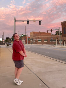 Logan Byrd stands on the sidewalk of a small-town street, smiling at the camera with his hands folded in front of him. He wears a dark red T-shirt, gray shorts, and white sneakers. A stoplight and crosswalk are visible in the background, along with a beautiful purple and pink sunset.