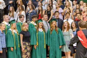 A choir sings the national anthem in a packed gymnasium during a graduation ceremony, with choir members standing in two rows and their director facing them with his back to the camera. The bleachers behind the choir are filled with spectators, many with their hands over their hearts. Logan Byrd stands on the far left of the back row, wearing green graduation regalia with yellow and white accents. Of the 14 visible choir members, six are wearing graduation regalia.