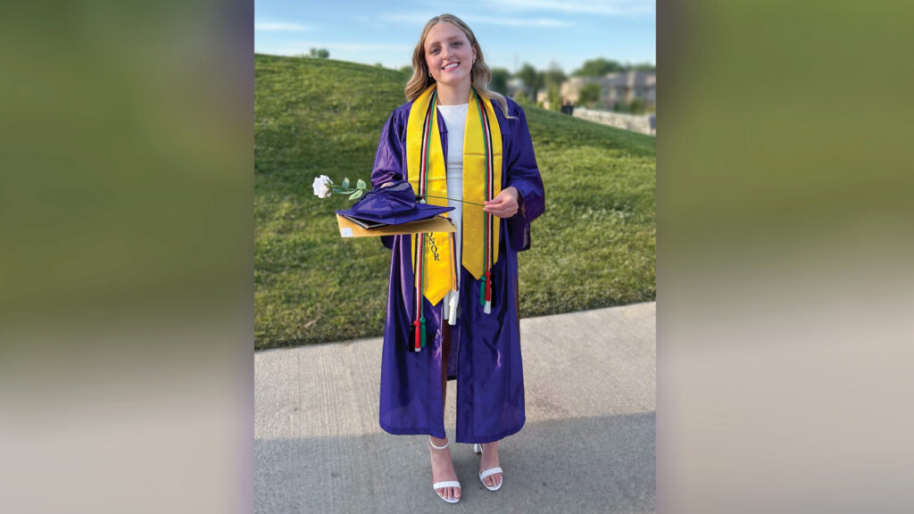 Nadia Cline stands in her high school graduation regalia on a sidewalk in front of a grassy hill, smiling at the camera. Her long, wavy blond hair falls loose past her shoulders. She wears a deep purple gown with a yellow stole embroidered with the word “HONOR” in capital letters, along with black, red, green, and white cords. She holds her graduation cap, diploma cover, a large manila envelope, and a white rose. Her unzipped gown reveals a knee-length white dress, and she wears white sandals with block heels.