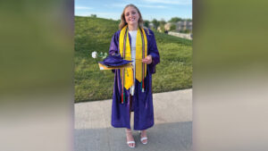 Nadia Cline stands in her high school graduation regalia on a sidewalk in front of a grassy hill, smiling at the camera. Her long, wavy blond hair falls loose past her shoulders. She wears a deep purple gown with a yellow stole embroidered with the word “HONOR” in capital letters, along with black, red, green, and white cords. She holds her graduation cap, diploma cover, a large manila envelope, and a white rose. Her unzipped gown reveals a knee-length white dress, and she wears white sandals with block heels.