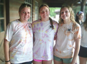 Three high school-aged young women stand together in a row with their arms around one another, smiling at the camera. They each wear a tie-dyed T-shirt with shorts. The shirts are slightly different colors and patterns, suggesting they may be handmade, and each features a 4-H logo with text surrounding it and additional text below on the left side. Behind them is a cabin-like building surrounded by trees, with a few other people nearby, giving the setting the appearance of a summer camp.
