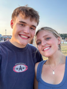 Nadia Cline stands closely beside a young man as they pose and smile at the camera, appearing to take a selfie together. Both wear blue: he wears a T-shirt with an Aéropostale logo, while she wears a blue ribbed tank top, silver necklace with a square charm, and silver earrings. Her long blond hair is pulled back into a braid. They are outdoors at what appears to be a county fairgrounds, with a large grassy field and a line of trees in the distance. People, portable toilets, and stands are visible in the background. The cloudless sky has begun to turn orange near the horizon as the sun sets.