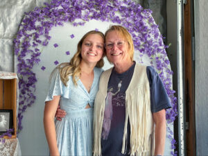 Nadia Cline stands closely beside an older woman with her arm around Nadia’s waist, their heads touching as they smile at the camera. The older woman has short blond hair and glasses and wears a white fringed vest over a T-shirt featuring Stevie Nicks’ Bella Donna album cover. Nadia wears a light blue short-sleeved dress with rows of flower-shaped eyelet detailing. They stand in front of a white arched backdrop decorated with purple flowers along the top. Nearby are lace-covered shelves displaying framed photographs, including a black-and-white photo of a young girl.