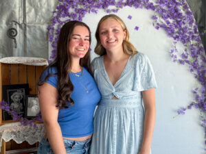 Nadia Cline stands closely beside a young woman with long dark hair, both smiling at the camera. The young woman wears jeans, a short blue T-shirt, and three layered necklaces. Nadia wears a light blue short-sleeved dress with rows of flower-shaped eyelet detailing. They stand in front of a white arched backdrop decorated with purple flowers along the top. Nearby, lace-covered shelves display framed photographs, including two black-and-white photos of a young girl. A vine of purple flowers from the backdrop extends along one of the shelves.