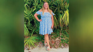 Reagan Bowers stands smiling on sand in front of dense tropical foliage. She poses with one foot crossed over the other and one hand on her hip. She wears a sky-blue, off-the-shoulder dress with a white daisy pattern. Her long blond hair is pulled back on the sides.