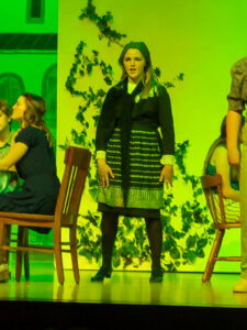 Reagan Bowers stands onstage singing in a theatrical production, between two tables surrounded by chairs occupied by other high school-age performers. A partially visible performer stands beside her. The set resembles an outdoor café, with a white wall covered in ivy, windows, and roof slats. Reagan wears a black-and-white long-sleeve blouse with a large collar, a knee-length black skirt, a black-and-white apron, black tights, and black shoes. A plain bandana covers her hair, and a black scarf or shawl is tied around her neck. Green stage lighting gives the entire image a green tint.