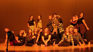 A large group of at least 18 high school-age performers pose onstage under strong orange lighting. Some sit while others stand behind them. They wear black shirts or tank tops, black leggings, and plaid button-up shirts either worn over their tops or tied around their waists. All are barefoot. The group appears to be holding a final pose at the end of a dance or performance, with most smiling toward the audience. Two performers hug in the back row. A light on a tripod stands at the front of the stage, illuminating the group. The background is a blank dark canvas, tinted orange by the stage lighting.