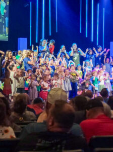 A large group of young children perform onstage, singing and making hand gestures as part of the song. Older high school-age performers stand among them, helping and interacting with the children. The children’s faces are slightly blurred to protect their identities. Several rows of adults sit in theater-style seating in the foreground, watching the performance. The stage backdrop is dark blue with two sets of cascading neon-blue vertical stripes. A portion of a large video screen showing a closer view of the performance is visible along the edge of the photo.