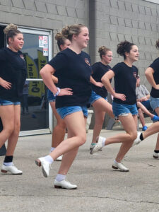 Reagan Bowers performs with the Hearts Ablaze clogging group in front of a gray brick building with a diamond pattern in the brickwork. Shown from the side, Reagan smiles as she looks ahead while dancing. Six dancers are partially or fully visible in two rows. All have their hair pulled back into large buns and wear denim shorts, black T-shirts featuring a red heart outline with white lettering, and white tap shoes. The dancers balance on their left feet with their right feet extended to the side in a dance move. A few people sit on a bench behind the dancers along the building’s wall.