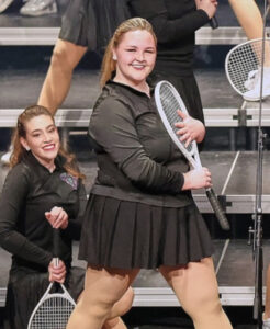 Reagan Bowers stands on risers onstage, smiling toward the audience while holding a tennis racket against her torso. She walks toward the right side of the photo while turning her head toward the audience. She and the other performers wear black tennis skirts, long-sleeve black polo shirts, and white tennis shoes, with each holding a tennis racket. Another girl kneels behind Reagan, holding her racket toward the floor while smiling at the audience. Both girls wear their hair in ponytails.