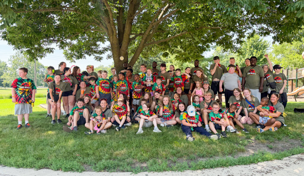 A group photo of Camp Achieve campers and staff posing beneath a tree at the edge of the Burris playground. Although it is a bright, sunny day, the group is shaded by the tree. Campers wear red, green, yellow, and black tie-dyed shirts featuring the Camp Achieve Jurassic Park logo, while staff wear olive green shirts with the same logo. Everyone is arranged in several rows, with some sitting on the grass and others standing. Some look at the camera and smile, while others look to the side or interact with one another.