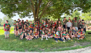 A group photo of Camp Achieve campers and staff posing beneath a tree at the edge of the Burris playground. Although it is a bright, sunny day, the group is shaded by the tree. Campers wear red, green, yellow, and black tie-dyed shirts featuring the Camp Achieve Jurassic Park logo, while staff wear olive green shirts with the same logo. Everyone is arranged in several rows, with some sitting on the grass and others standing. Some look at the camera and smile, while others look to the side or interact with one another.