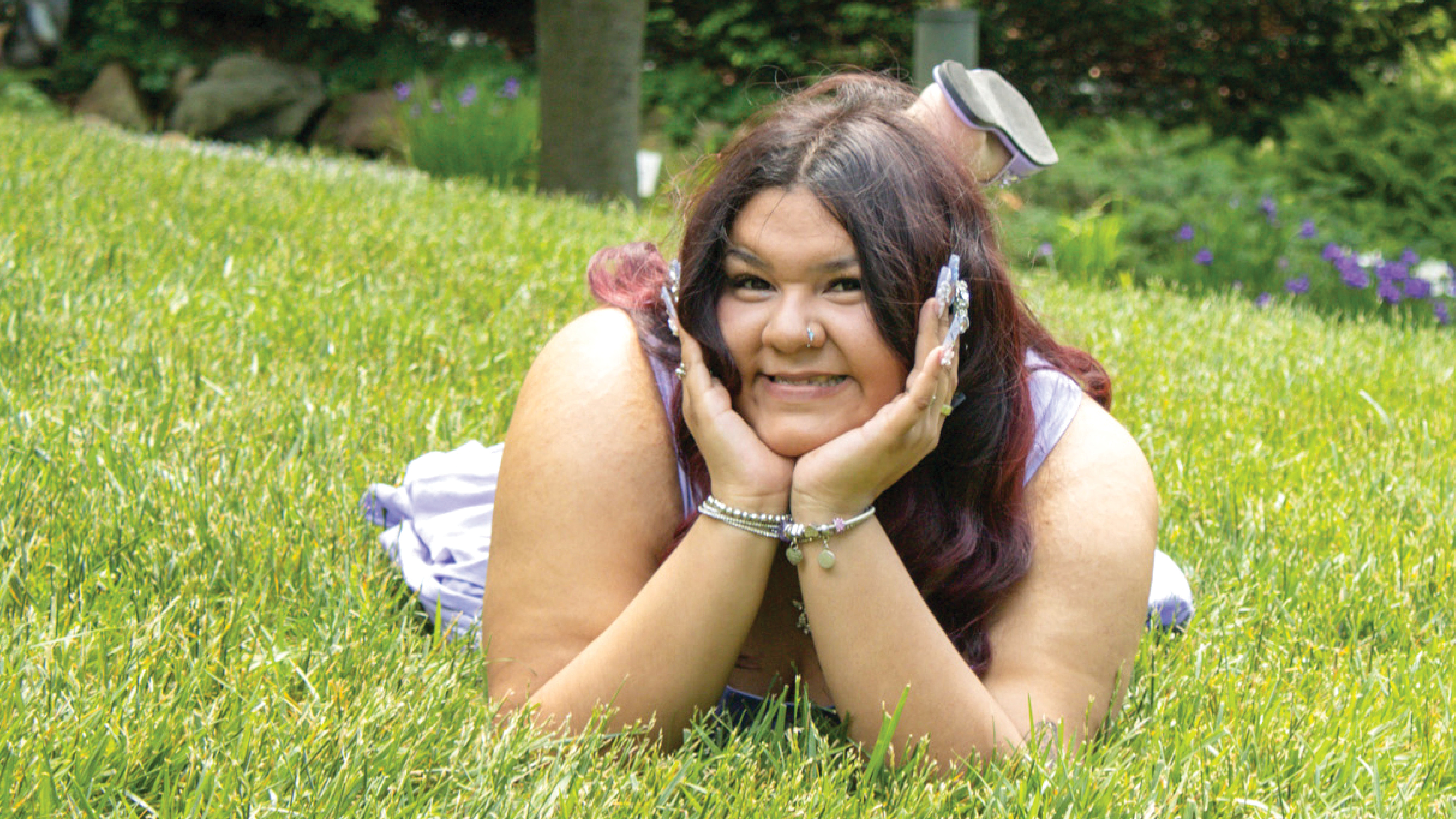 Damariz Alvarado lays on a grass covered soft hill posing in a traditional senior picture pose with her elbows on the ground, and her head propped up by her hands. Her chin is situated where her two hands meet, and her fingers are on either side of her face, pointing upwards. You can see one of her feet behind her head, as she has at least one knee bent. She is wearing sandals. She is wearing pale purple, silver bracelets, and her long, decorated fingernails have jewels on them. In the background you can see where the edge of the lawn meets some shrubs and a tree. There are flowers, a birdhouse or bird feeder that looks like a gazebo, and a statue of a figure.