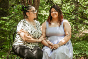 Damariz Alvarado sits beside an older woman, likely a family member, in a forested area. The pair smile as they sit with their arms intertwined and hold hands in Damariz’s lap. The older woman looks toward Damariz, while Damariz looks at the camera. Sunlight filters through the trees, creating bright spots against the muted forest tones. Damariz has long, slightly curly brown hair that transitions to red at the ends and wears a pale gray or purple dress. The older woman wears black jeans and a white blouse with a black animal-print pattern.