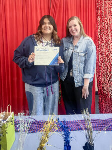 Damariz Alvarado poses with a woman in front of a bright red curtain, with a red beaded curtain visible on the right. Damariz wears graduation tassels with jeans, a T-shirt, and a partially zipped navy blue sweatshirt. A pile of additional red and blue tassels sits on a table in front of them, along with metallic party favors and a green gift bag. Damariz holds a paper that reads “Certificate of Mastery.”