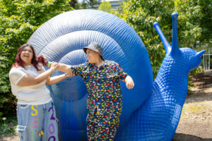 Damariz Alvarado and another person stand in front of a large blue snail sculpture that towers over them. Damariz, on the left, wears wide-leg jeans with lettering painted on the legs, possibly reading “Senior” and “2026.” Both are laughing and smiling rather than looking at the camera, as the other person playfully holds Damariz’s hair. The other person wears a matching black button-up shirt and pants covered in brightly colored dinosaur patterns, along with a coordinating bucket hat decorated with colorful dots. They are outdoors on a sunny day, with trees and bushes visible behind the sculpture.