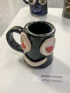 A handmade mug sits on a white table in front of two partially visible sculptures. A tag in front of the mug identifies Damariz Alvarado as its maker and lists Applied Ceramics as the class in which it was created. The mug is shiny black with a matching black handle. A face is applied to the side, featuring large white circular eyes with red heart-shaped pupils and a simple, curved smile.