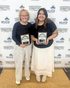 Damariz Alvarado and an older woman stand smiling at the camera in front of a white backdrop featuring the Decatur Township logo and the words “Decatur Proud” repeated across it. Both wear black tops with white bottoms. The older woman wears a long-sleeved top with white slacks featuring thin black stripes, while Damariz wears a short-sleeved top and a long white skirt with lace along the bottom. Both hold plaques that feature “Decatur Central High School” in an arch across the top, a stylized intertwined “D” and “C” surrounded by wings, and possibly the word “Banquet.”