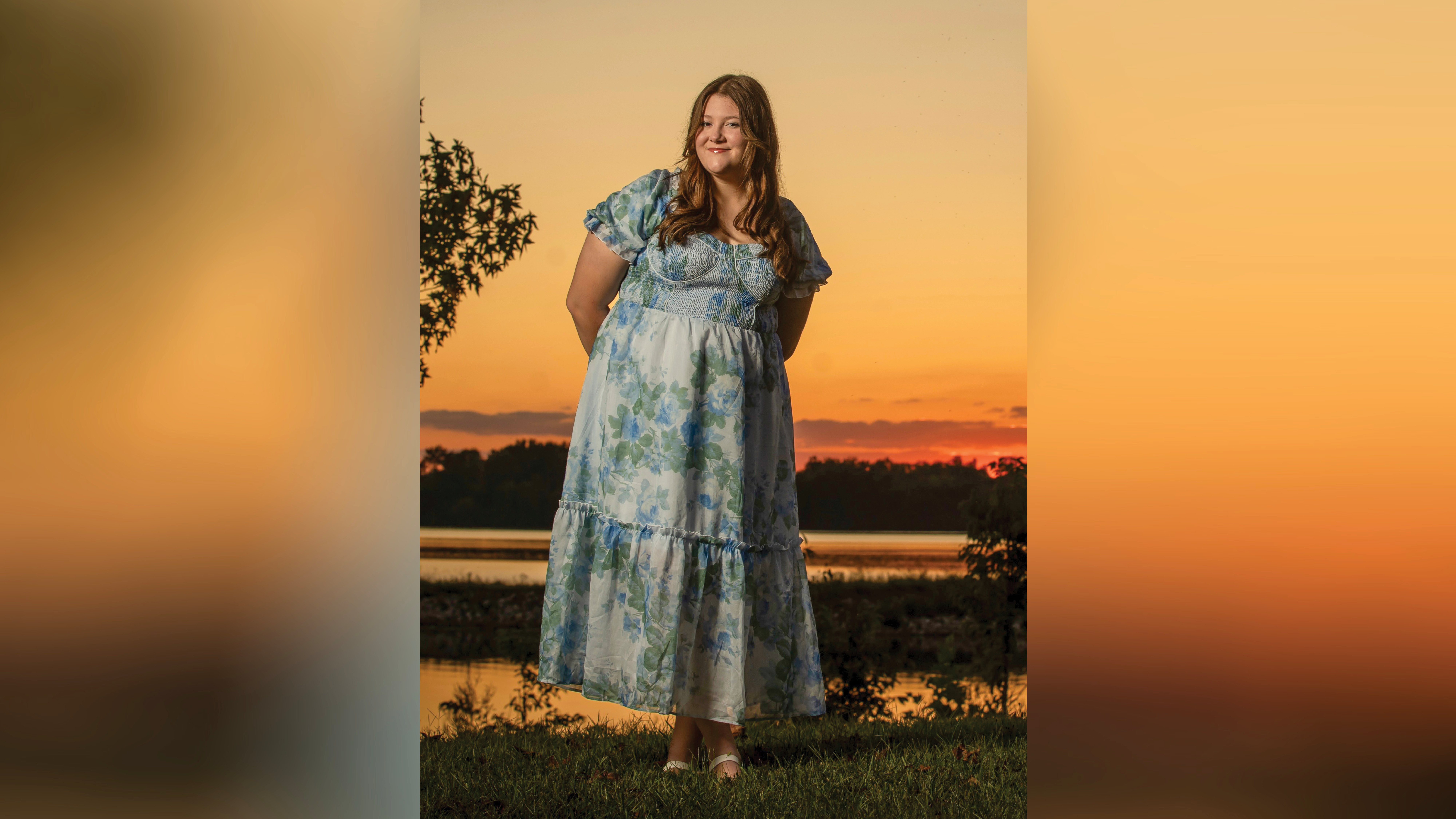 Kylee VanMeter smiles at the camera while standing in front of a small lake at sunset. The orange sky and sunset reflect on the water, with a dark row of trees along the opposite shore. She stands with one foot crossed over the other and her hands behind her back. She wears a blue floral tiered dress with white sandals, and her long brown hair falls past her shoulders.