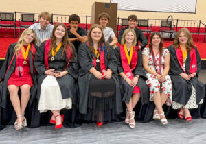 Kylee VanMeter sits among a group of peers at what appears to be a high school graduation ceremony. She is one of six young women seated on the edge of a stage, five of whom wear graduation gowns with red stoles and gold Academic Honors Medallions featuring a blue Indiana state emblem. Another young woman wears two honor cords but no gown. Four young men stand behind them. The stage is set with chairs and a podium against a tan brick wall with a screen, suggesting a high school gymnasium.