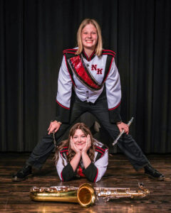 Two high school-age young women pose on a polished wood stage with a black curtain backdrop. Kylee VanMeter lies on her stomach facing the camera, propping her head in her hands with one foot raised behind her. A tenor saxophone rests on the floor in front of her. Her friend stands over her with a foot on either side, holding a flute and piccolo while resting her hands on her knees. Both smile at the camera and wear black pants with gray marching band jackets featuring red and black accents and a stylized “NK” on the front. Kylee has long brown hair, while her friend has shoulder-length blonde hair.