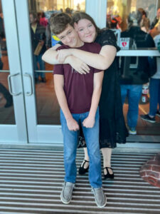 Kylee VanMeter hugs a younger boy while posing for the camera in front of glass and silver doors leading into a crowded event space. She rests her head against his as she wraps her arms around him, while he smiles with his hands held in front of him. Kylee wears a black lace dress and black dress shoes, while the boy wears jeans and a maroon T-shirt.