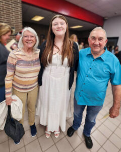 Kylee VanMeter poses with an older woman and man in a crowded school hallway, likely around the time of her high school graduation. The older woman and man each have an arm around Kylee as they smile for the camera. Kylee wears a white dress, white sandals, and a black cardigan, with her long brown hair worn loose. The older woman wears khaki pants and a striped long-sleeve shirt and holds a white jacket and black purse. The older man wears jeans and a teal short-sleeve button-up shirt. Both have white hair.