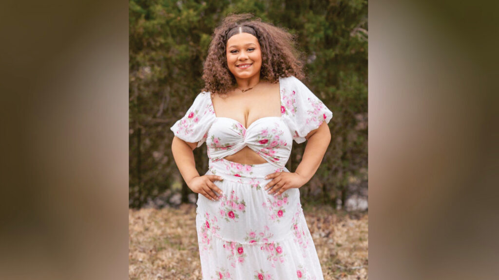 Callie Williams stands outdoors in front of a row of evergreen trees, smiling at the camera with her hands on her hips. Her shoulder-length curly brown hair is blowing in the wind. She wears a white tiered dress with a pink and magenta floral pattern, flowy sleeves, a twisted fabric detail on the bodice, and a midriff cutout. She also wears a necklace with cursive lettering.