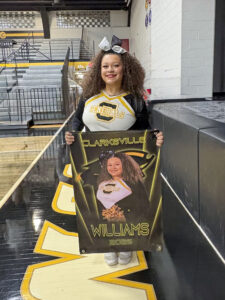 Callie Williams stands in a high school gym holding a large poster featuring a photograph of herself in her cheerleading uniform. Williams has curly brown hair pulled into a ponytail with a large black-and-white bow and is smiling while wearing red lipstick. She wears a black, white, and yellow long-sleeve cheerleading uniform. In the poster photo, she wears the same uniform with her hair down and holds black and gold pom poms in front of a stylized background featuring her school logo. The poster reads “Clarksville,” “Williams,” and “2026.” A stage is visible next to her.