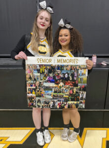 Callie Williams and a friend stand in a high school gym holding a large poster covered with photographs. The poster reads “Senior Memories,” with “2026” displayed vertically between the two words. Both women wear black, white, and yellow long-sleeve cheerleading uniforms, with their hair pulled into high ponytails and large black-and-white bows. Callie has shoulder-length curly hair, while her friend has long, straight blond hair. Both are wearing red lipstick. A black stage with its curtain closed is visible behind them.