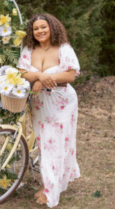 Callie Williams stands outdoors on brown grass in front of a row of evergreen trees, smiling at the camera with her hands resting on the handlebars of a pale yellow vintage bicycle. A basket filled with white daisies and yellow daffodils is attached to the handlebars, with more of the same flowers decorating a nearby metal garden feature. Williams has shoulder-length curly brown hair and wears a white tiered dress with a pink and magenta floral pattern, flowy sleeves, a twisted fabric detail on the bodice, and a midriff cutout. She also wears a necklace with cursive lettering and is barefoot.