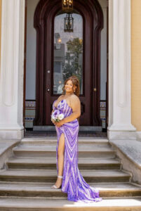 Callie Williams poses on the front steps of a large, stately house with tall pillars and an oversized dark wood and glass door. She wears a fitted purple and white prom dress with a swirling pearl and sequin pattern and a side slit. Her hair is styled in an updo with a pearl accent and curled tendrils framing her face. She holds a small bouquet of purple and white flowers and wears a sparkly, strappy high heel visible through the dress slit.