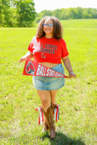 Callie Williams stands in a grassy field on a sunny day with a row of trees in the background. She wears a red Ball State Cardinals crop top featuring the Ball State athletic logo, a denim mini skirt, and brown cowboy boots decorated with red, white, and blue ribbons. She holds a red pennant reading “Ball State Cardinals” along the side and featuring a large Charlie Cardinal athletic logo. She has shoulder-length curly brown hair and wears tortoiseshell glasses.