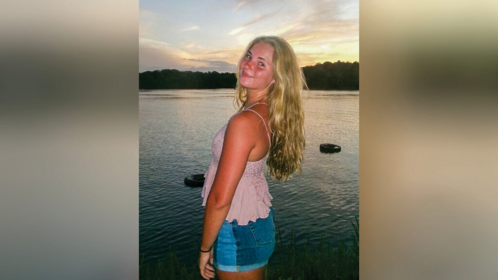 Addie Rosebrock stands beside a lake at sunset, facing left while looking back toward the camera with a soft smile. She wears a dusty pink tank top and blue jean shorts. Two tires float in the water, with a line of trees visible across the lake.