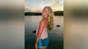 Addie Rosebrock stands beside a lake at sunset, facing left while looking back toward the camera with a soft smile. She wears a dusty pink tank top and blue jean shorts. Two tires float in the water, with a line of trees visible across the lake.
