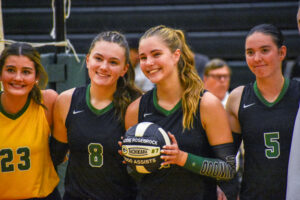 Four volleyball players pose together on an indoor volleyball court. Addie Rosebrock holds a volleyball commemorating her 1,000 assists, with her name and jersey number, #7, displayed on the ball. Three players wear black jerseys with green accents, while one wears a yellow jersey with green accents.
