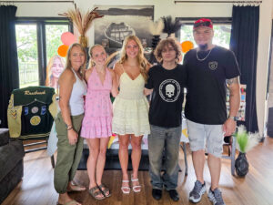 Five people stand together in a line with their arms around one another, smiling at the camera. Addie Rosebrock stands in the center wearing a yellow sundress and white sandals. Balloons, a large photo of Addie, and her letterman jacket are displayed in the room, suggesting the photo was taken at a graduation celebration. Large windows and a scenic black-and-white lake photograph are visible in the background.