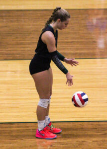 Addie Rosebrock prepares to serve during a volleyball game in a gym. A red, white, and blue volleyball hovers in the air after she bounced it on the floor. She wears a black uniform with dark green compression sleeves reading “GREENWOOD,” knee pads, and bright pink sneakers.