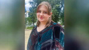 Evvy Kikkert poses for a profile photo outdoors, smiling softly. She wears a patterned teal, brown, and black shirt with a silver necklace and earrings. Trees, buildings, a statue, and street lights are visible in the background.