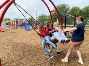 Evvy Kikkert pulls back a large inner-tube swing carrying several children on a playground, preparing to release it. The children are smiling and laughing, with their faces blurred to protect their identities. Other children play on equipment in the background.
