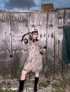 Evvy Kikkert poses in a blood-stained nurse costume while working at a haunted house. She wears a brown knee-length dress, black boots, a blood-covered nurse’s cap, and dark makeup, and holds a prop blade toward the camera. She stands in an alley in front of a tall wooden wall beneath dark, cloudy skies.