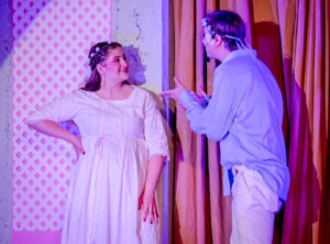 Evvy Kikkert performs onstage with a young man who gestures toward her as they appear to converse. She smiles at him with one hand on her hip. She wears a light-colored dress, while he wears a blue button-down shirt and tan pants. Purple stage lighting casts a purple hue over the scene. A lattice-covered stage wall and yellow curtain are visible behind them.