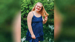 Lillyana Ray smiles at the camera while posing beside a pond. She wears a short blue sundress with a ruffled layer, and her blonde hair is pulled back on the sides. A line of trees is visible across the pond.