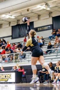 Lillyana Ray jumps to serve a volleyball during a high school game, her right arm extended as the black-and-white ball hovers above her. She wears a black and gold uniform, white kneepads, and white sneakers. Coaches and teammates watch from the bench, with spectators in the bleachers and game officials or announcers at a nearby table.