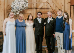 Seven people pose in a row at a wedding, with their arms around one another. Lillyana Ray stands on the far left in a pale blue dress. The bride wears a white wedding dress, while the others wear shades of blue. A large blue-and-white floral decoration hangs behind the group in front of a wooden door.