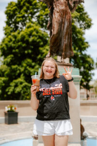 Lillyana Ray smiles in front of the Beneficence statue on Ball State’s campus, making the Chirp! Chirp! hand gesture. She wears a Ball State Cardinals shirt and white shorts and holds an iced coffee cup. A leafy tree is visible in the background on the sunny day.