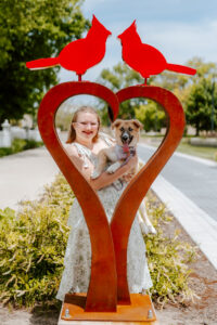 Lillyana Ray smiles at the camera while standing behind Ball State’s Love Birds sculpture, with her and her tan-and-white dog framed within the metal heart. She wears a white sundress. Two bright red cardinal figures sit atop the heart, with the Ball State campus visible in the sunny background.